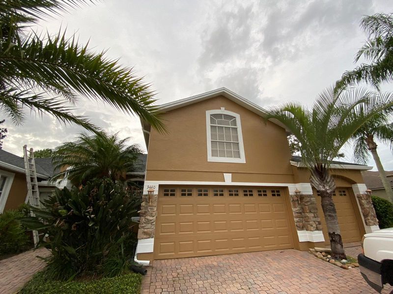 Tan house with brown garage doors, stone accents, palm trees, and overcast sky.