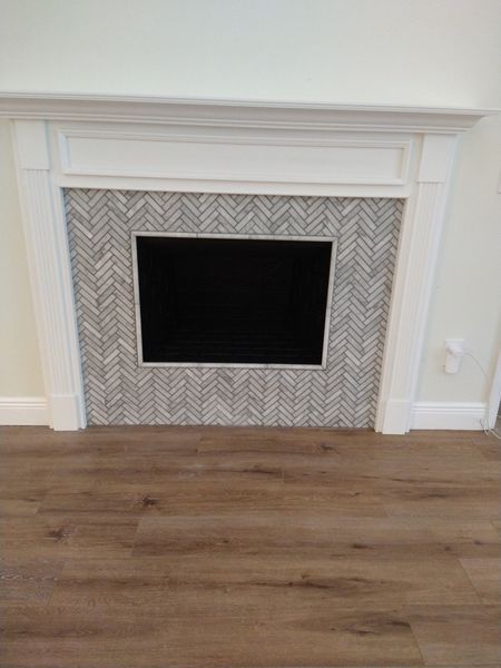 Fireplace with white mantel, grey herringbone tile, and black interior against light brown flooring.