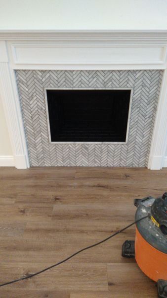 Fireplace with herringbone tile surround, dark opening, and white mantle. Flooring is wood grain.