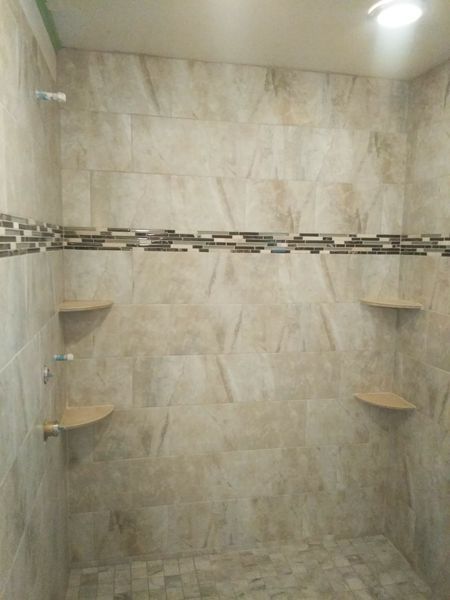 Tiled shower with neutral-toned walls, decorative trim, built-in corner shelves, and a ceiling light.