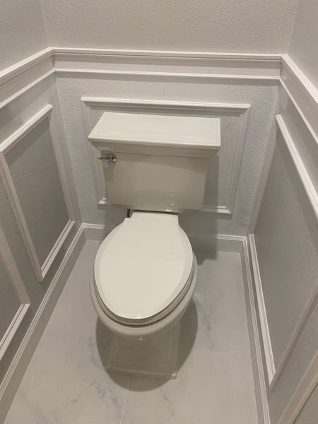 White toilet in a small bathroom with white molding on the walls and a white marble floor.