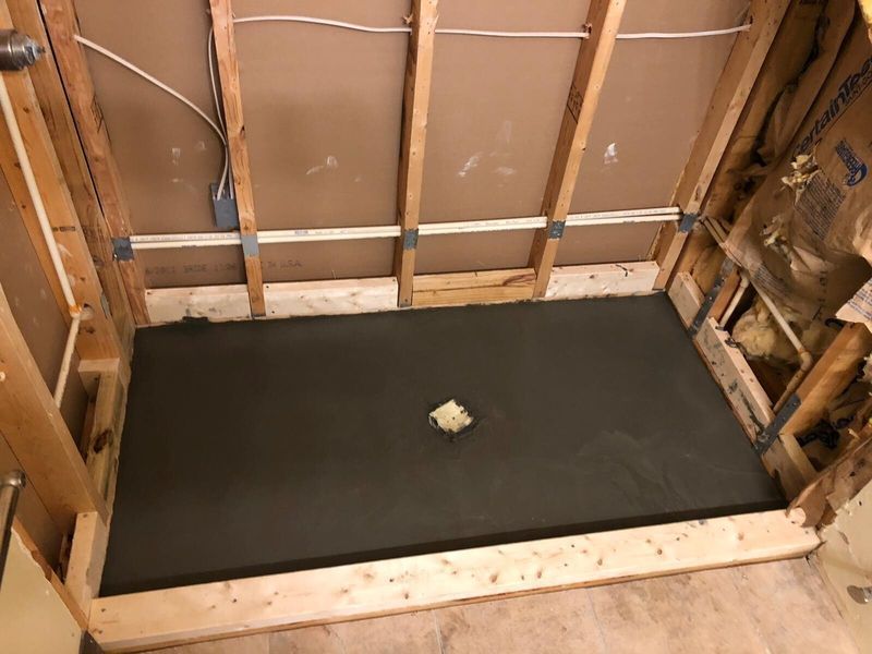 Shower base construction in progress, gray cement base set within a wooden frame, drain visible.