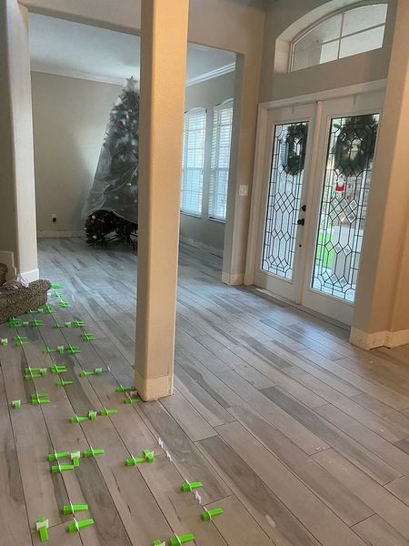 Tiled floor installation in a home. New gray tiles with green leveling spacers. Doors and a Christmas tree visible.