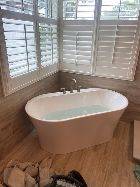 Freestanding white bathtub filled with water in a bathroom with window shutters and light wood tile flooring.
