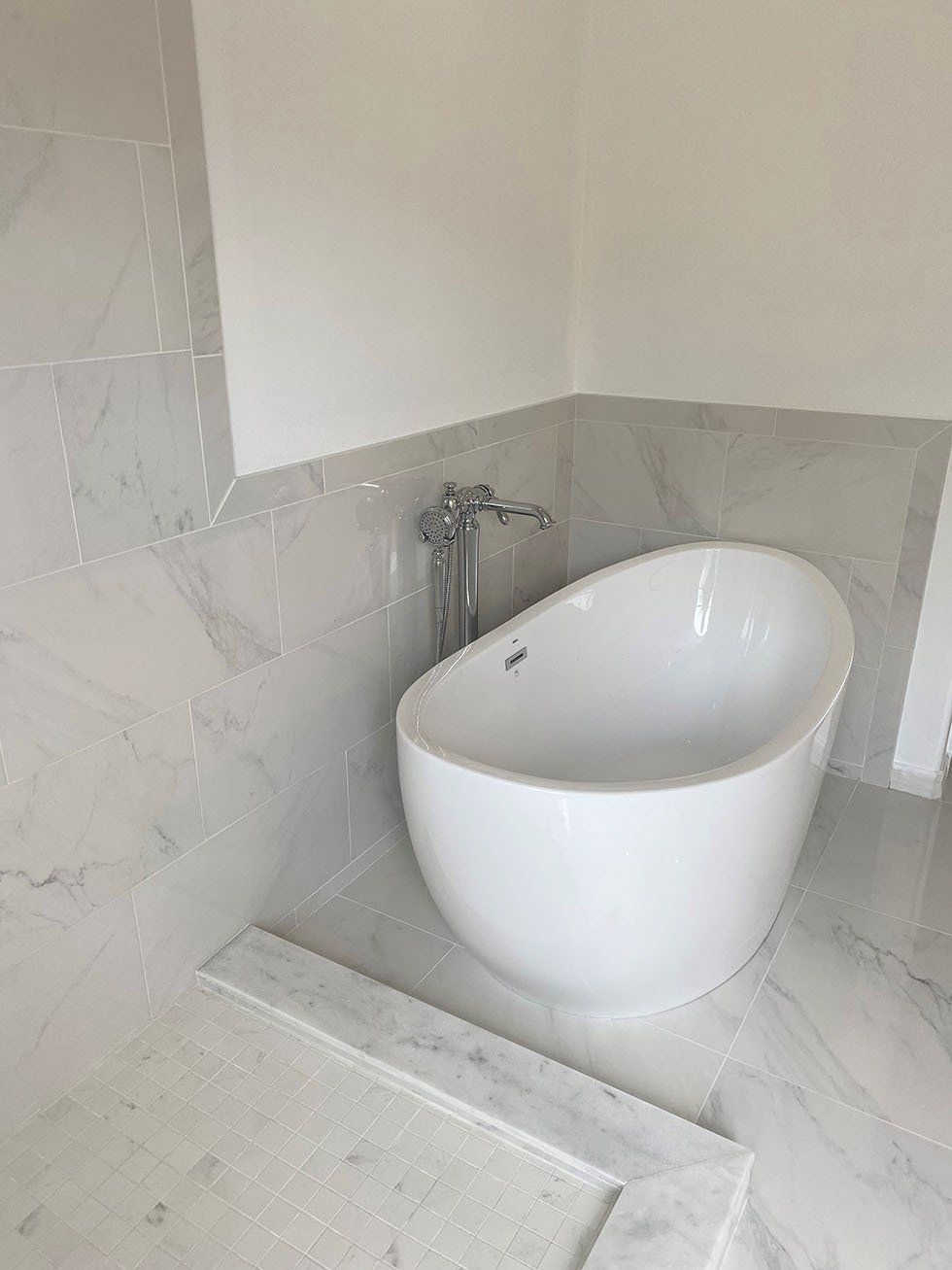 White oval bathtub with chrome faucet against marble tiled wall.