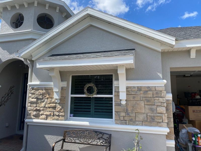 Gray stucco house with stone accent, window with green shutters and wreath, and decorative awning.