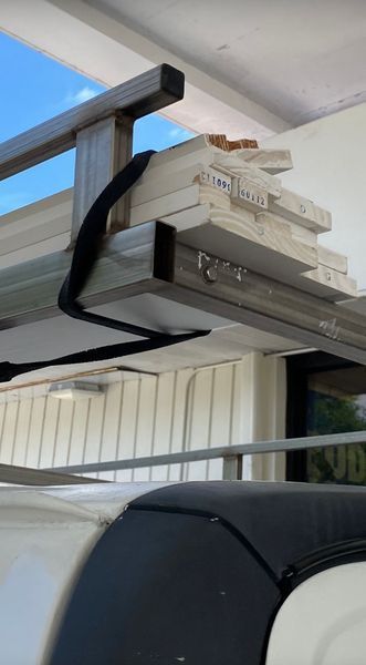 Wood planks secured to a roof rack with a black strap, mounted on a white vehicle.