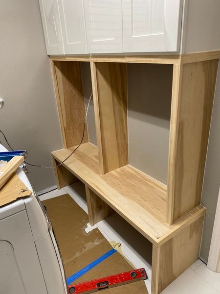 Light wood built-in shelving unit with white cabinets, being installed in a laundry room next to a white washing machine.