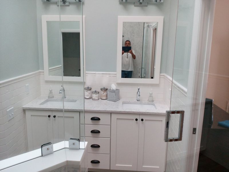 Bathroom with white cabinets, marble countertop, two sinks, mirrors, glass shower door, and a person taking a photo.