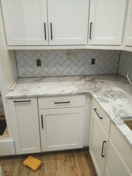 White kitchen cabinets with black hardware, marble countertop, and herringbone backsplash.