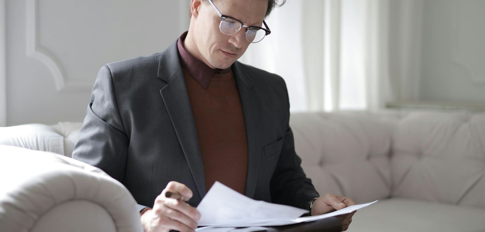 a man in a suit and glasses is sitting on a couch holding a piece of paper  in regards to signing up for health insurance. 