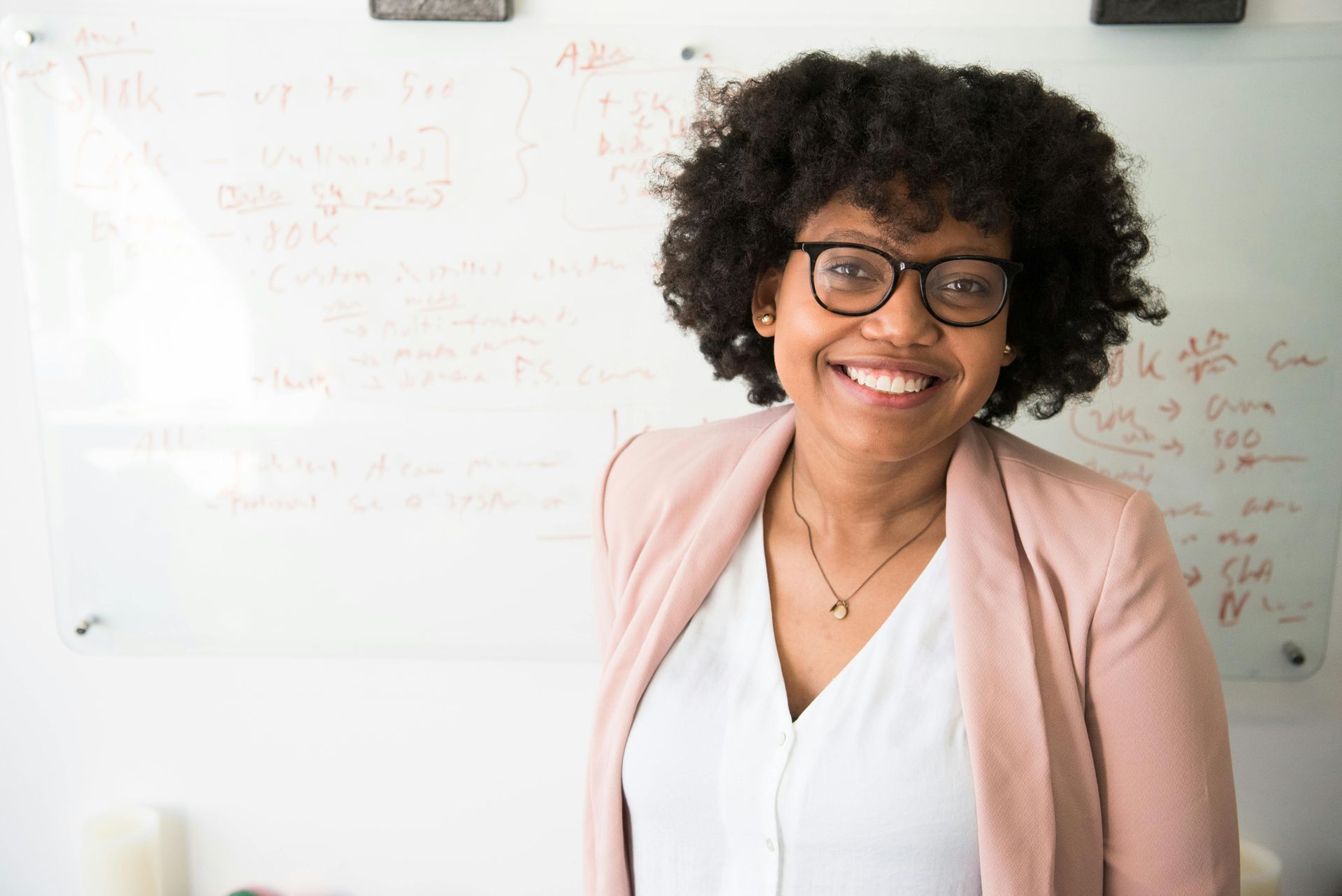 A woman wearing glasses is smiling in front of a whiteboard.