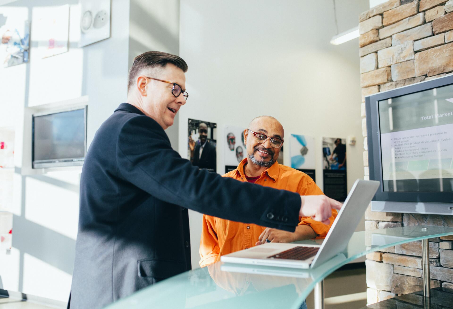 a man is pointing at a laptop computer while another man looks on .