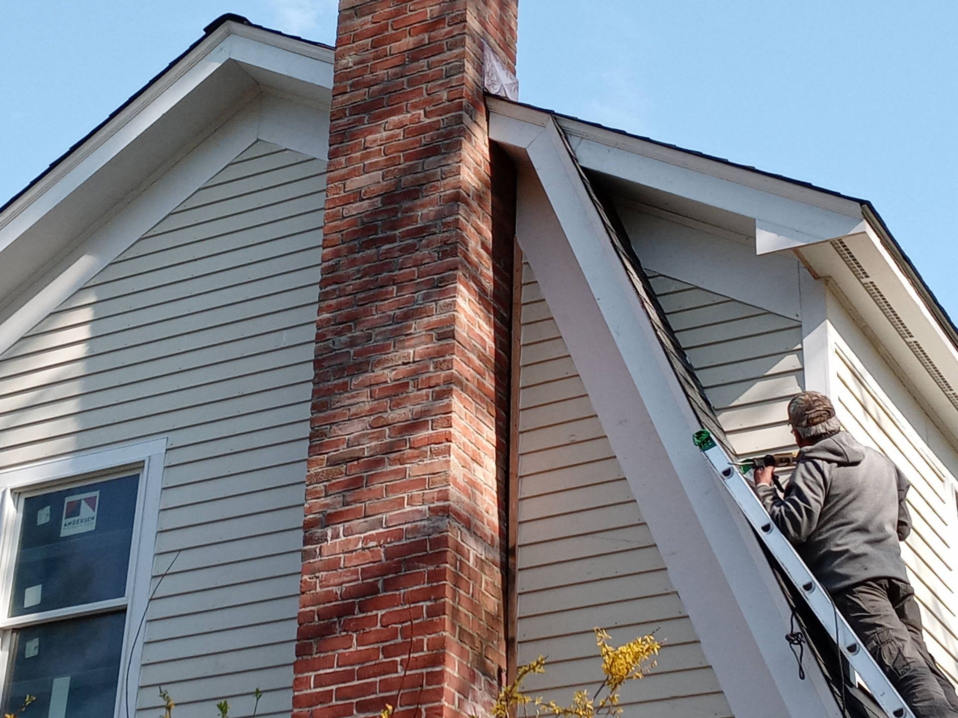 A man on a ladder is working on a chimney on the side of a house