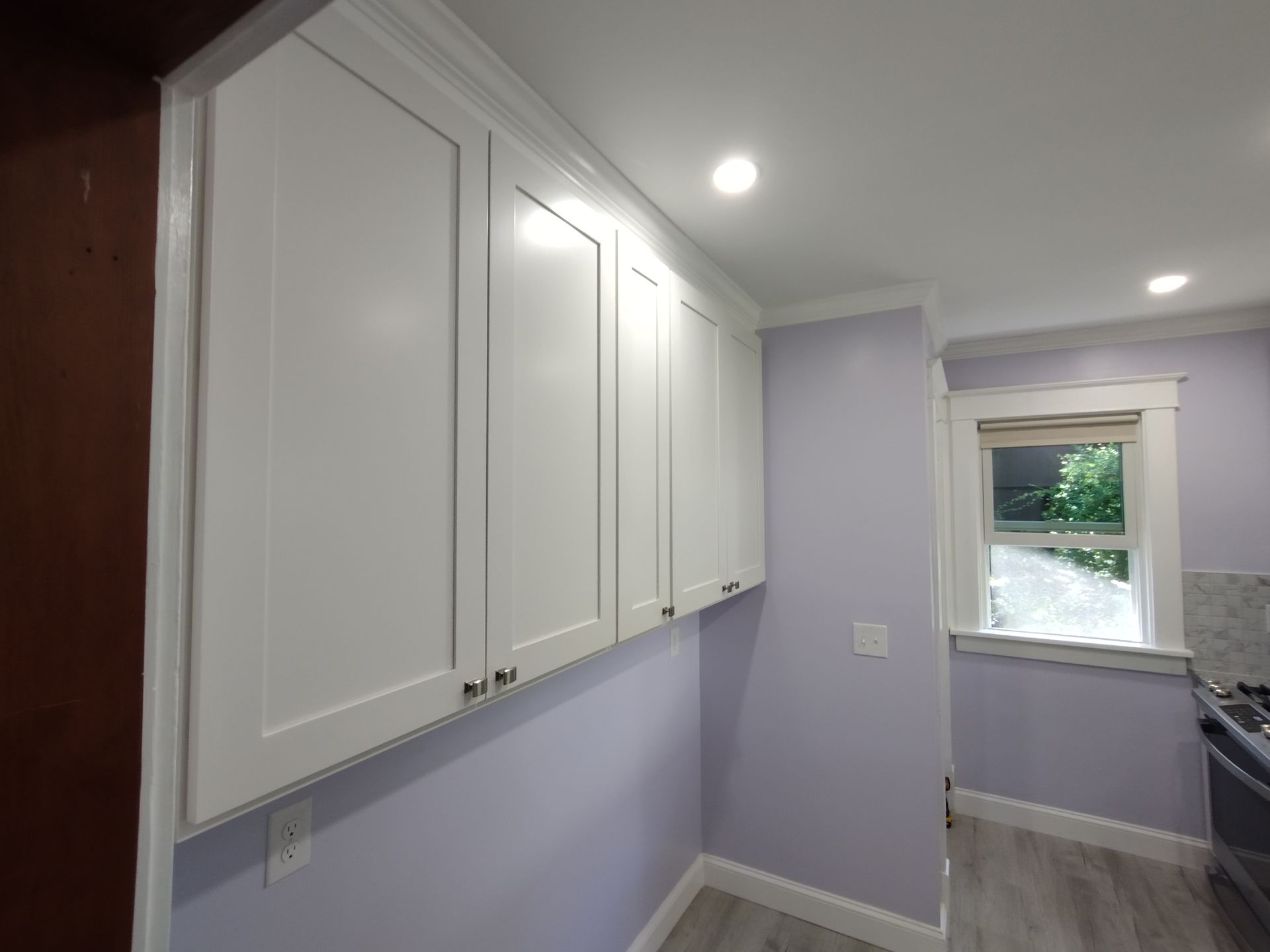 A kitchen with purple walls and white cabinets and a window.