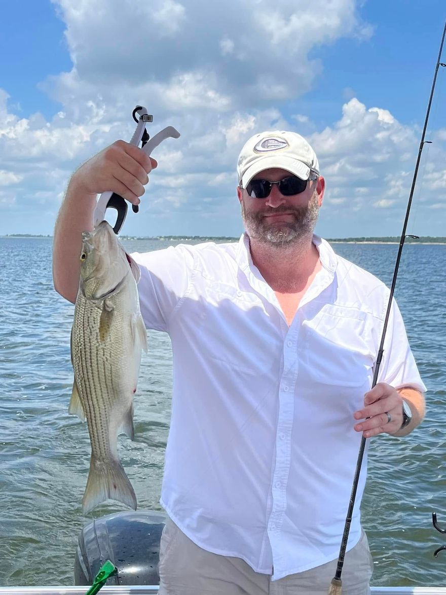 Man on a boat holding a fish with pliers, fishing rod, bright sky, water.