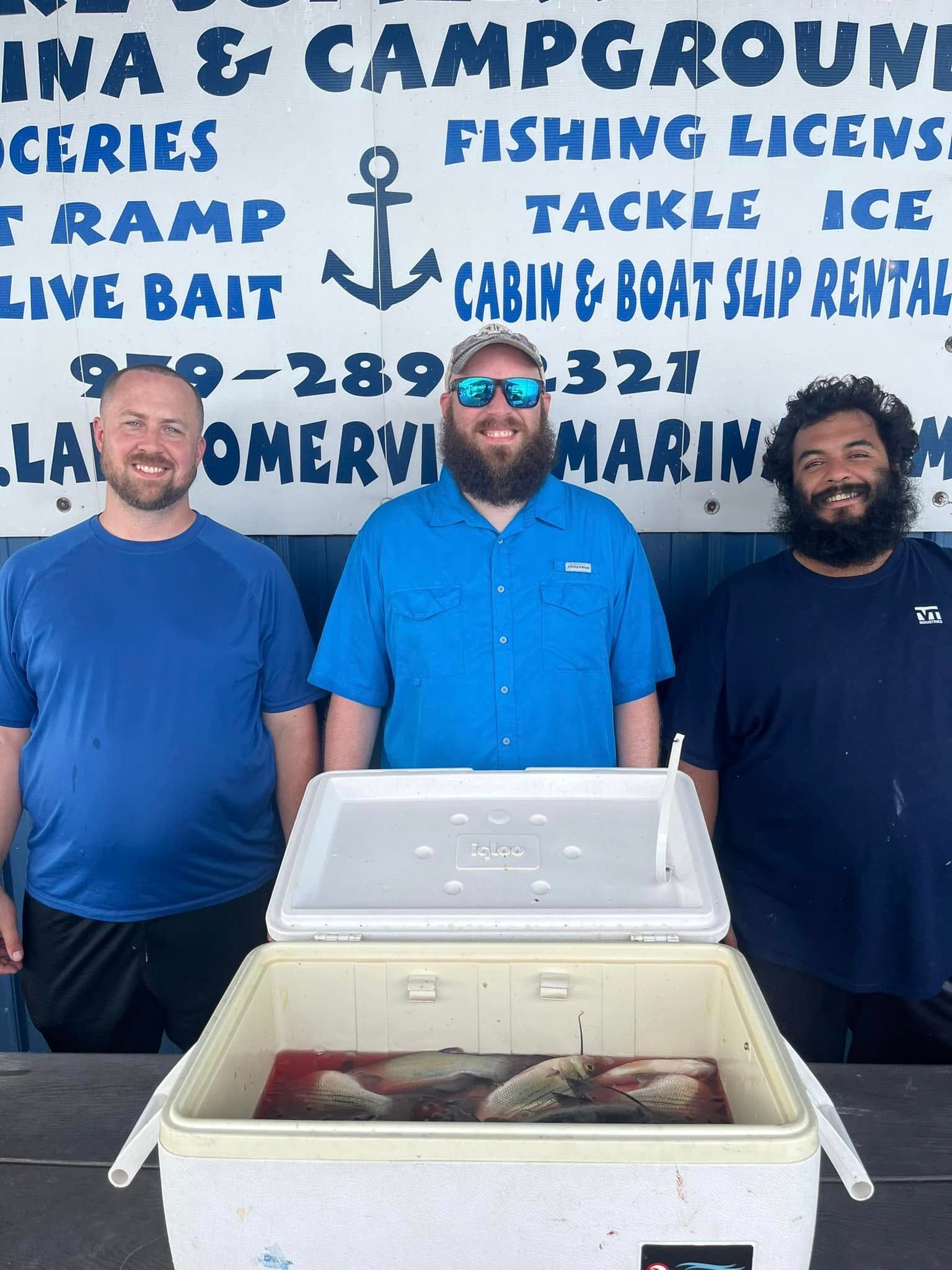 Three men stand in front of a marina sign with a cooler containing fish.