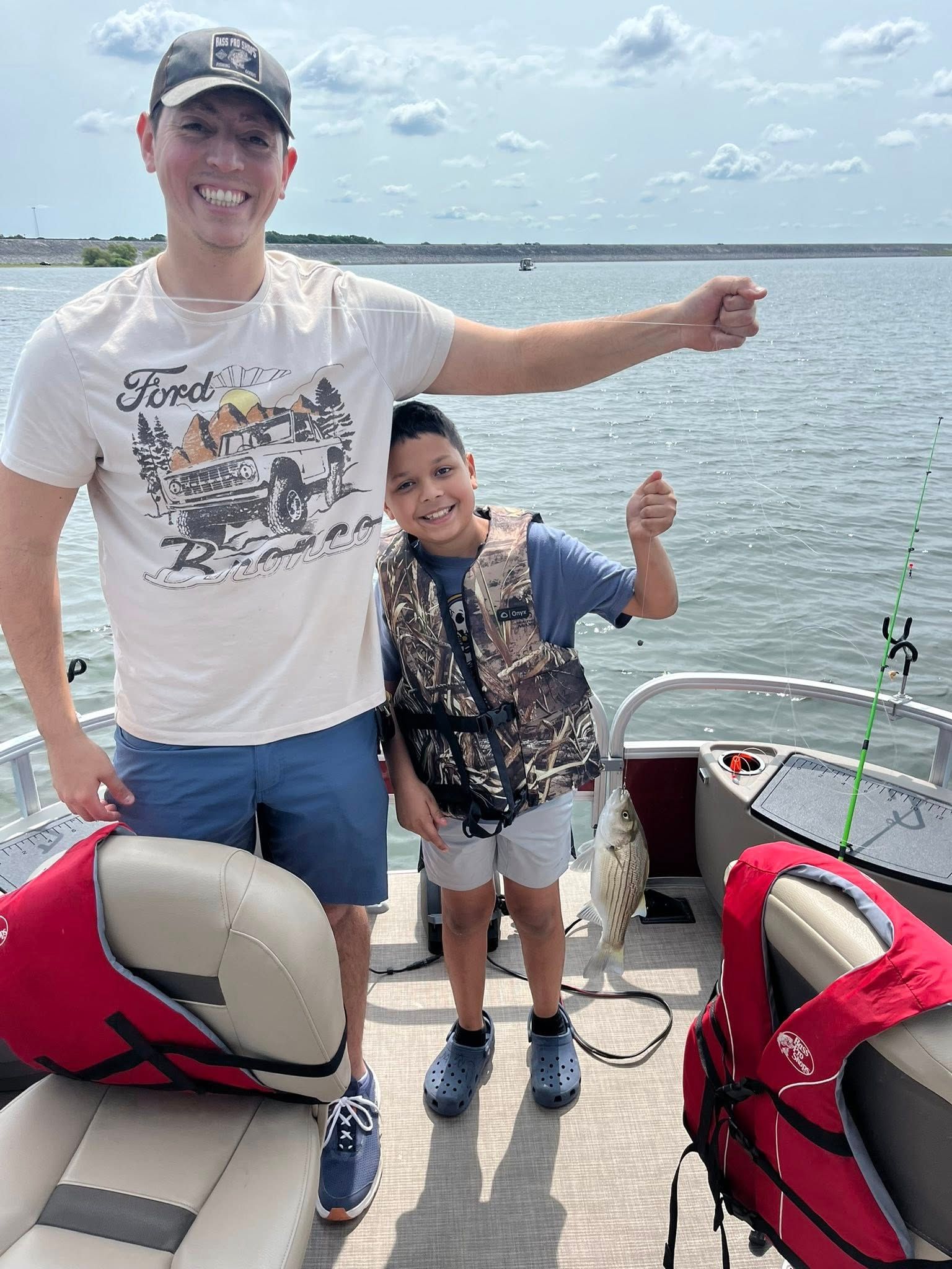 Man and child on a boat fishing. Child holds a fish, both smiling. Lake in background.