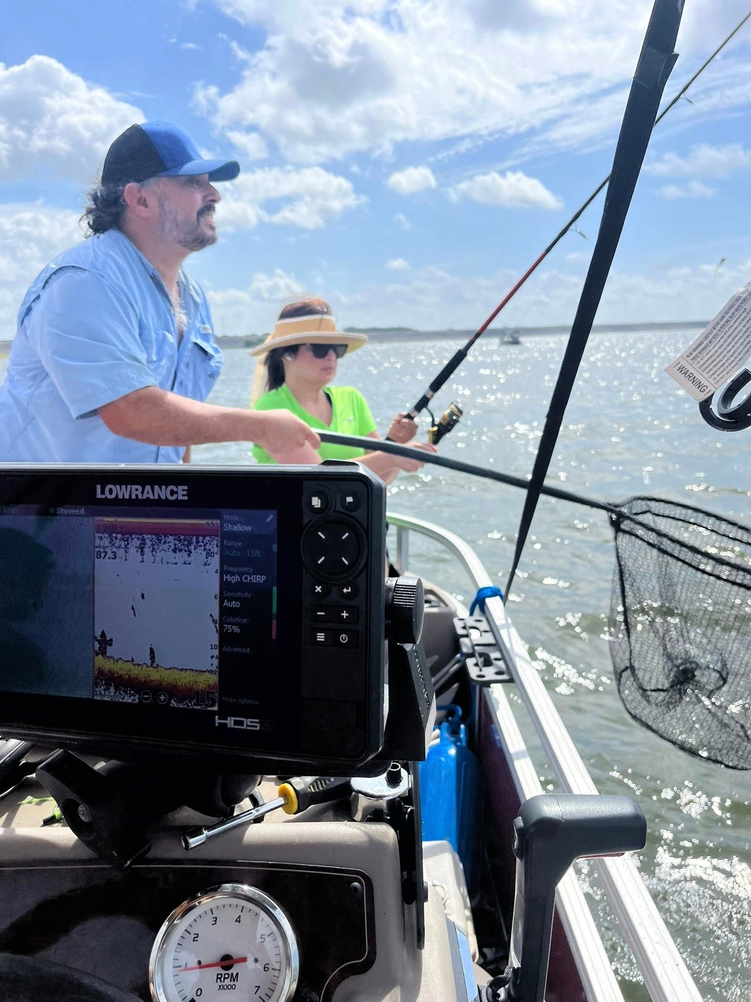 Man and woman fishing from a boat on a sunny day. The man wears a blue hat, and the woman wears a visor.