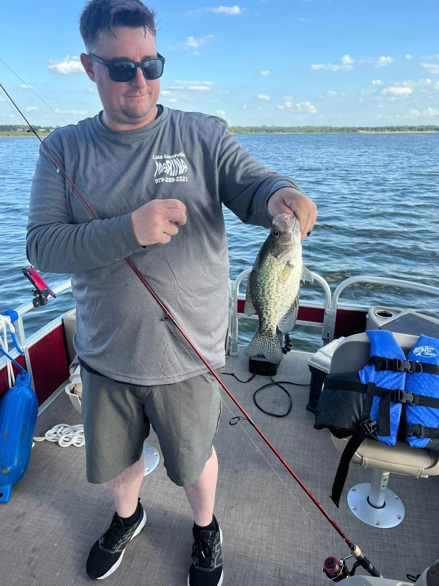 Man on a boat holding a fish he caught, smiling. Gray shirt, sunglasses, shorts, fishing rod. Outdoors, lake.