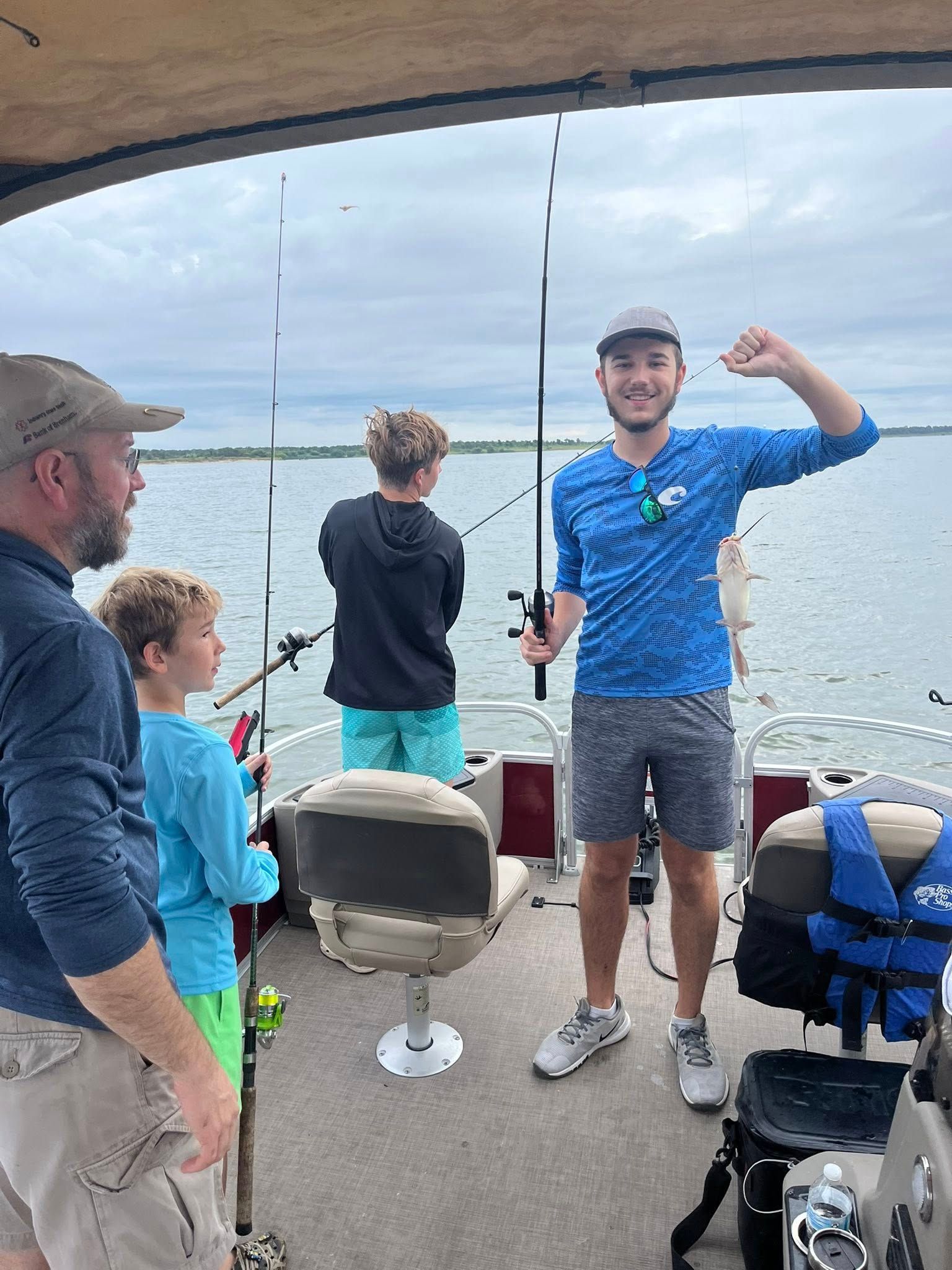 People fishing on a boat: man flexing, two boys, older man in a blue shirt, water background.