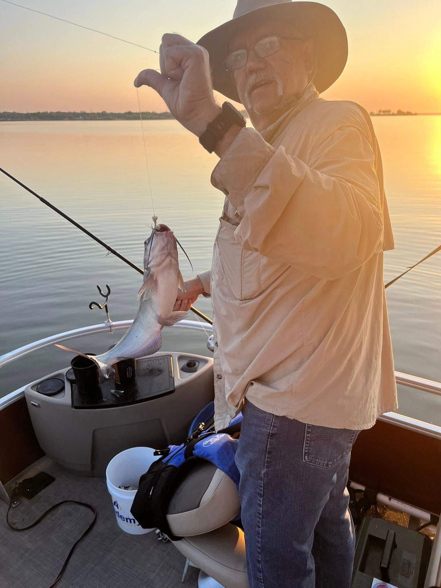 Man on a boat holds up a fish he caught, silhouetted by a sunset over calm water.