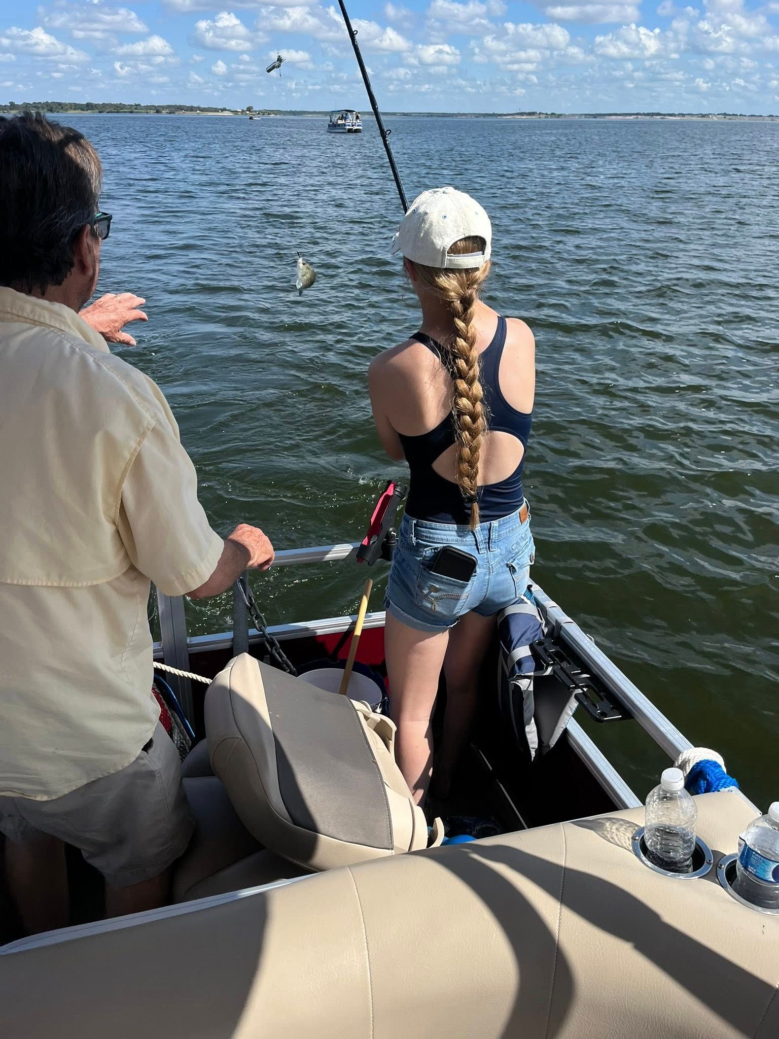 Woman fishing from a boat, holding a fishing rod. Man points, lake, sunny day.