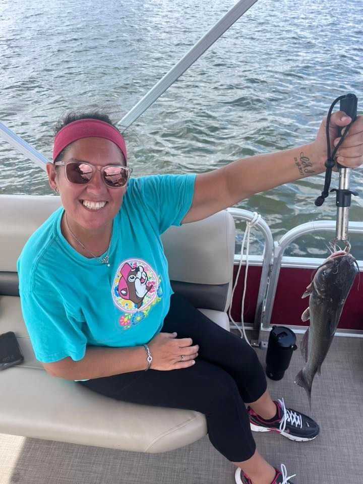 Woman on a boat smiles while holding a fish up by a scale. She wears sunglasses, blue shirt, and a red headband.