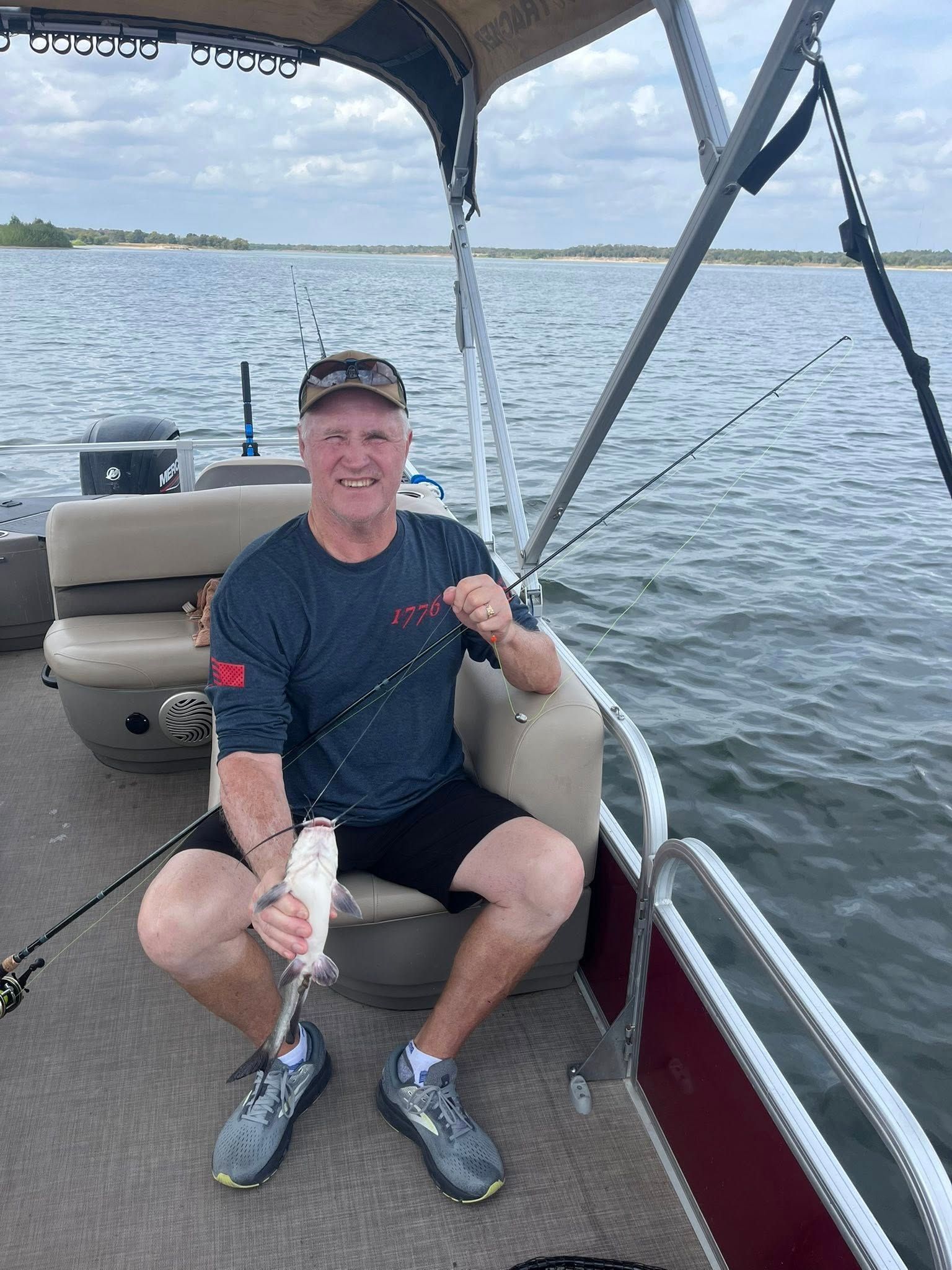 Man on a boat holds up a fish, smiling. He's wearing a hat, shirt, shorts. Cloudy sky, water in background.