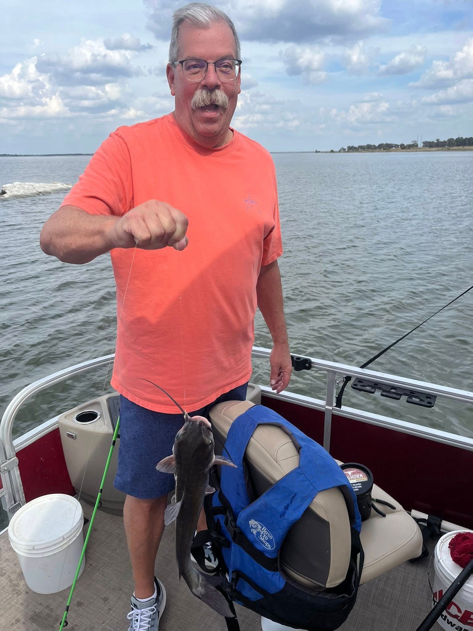 Man on a boat, holding up a catfish. He wears an orange shirt and blue shorts. Lake in background.