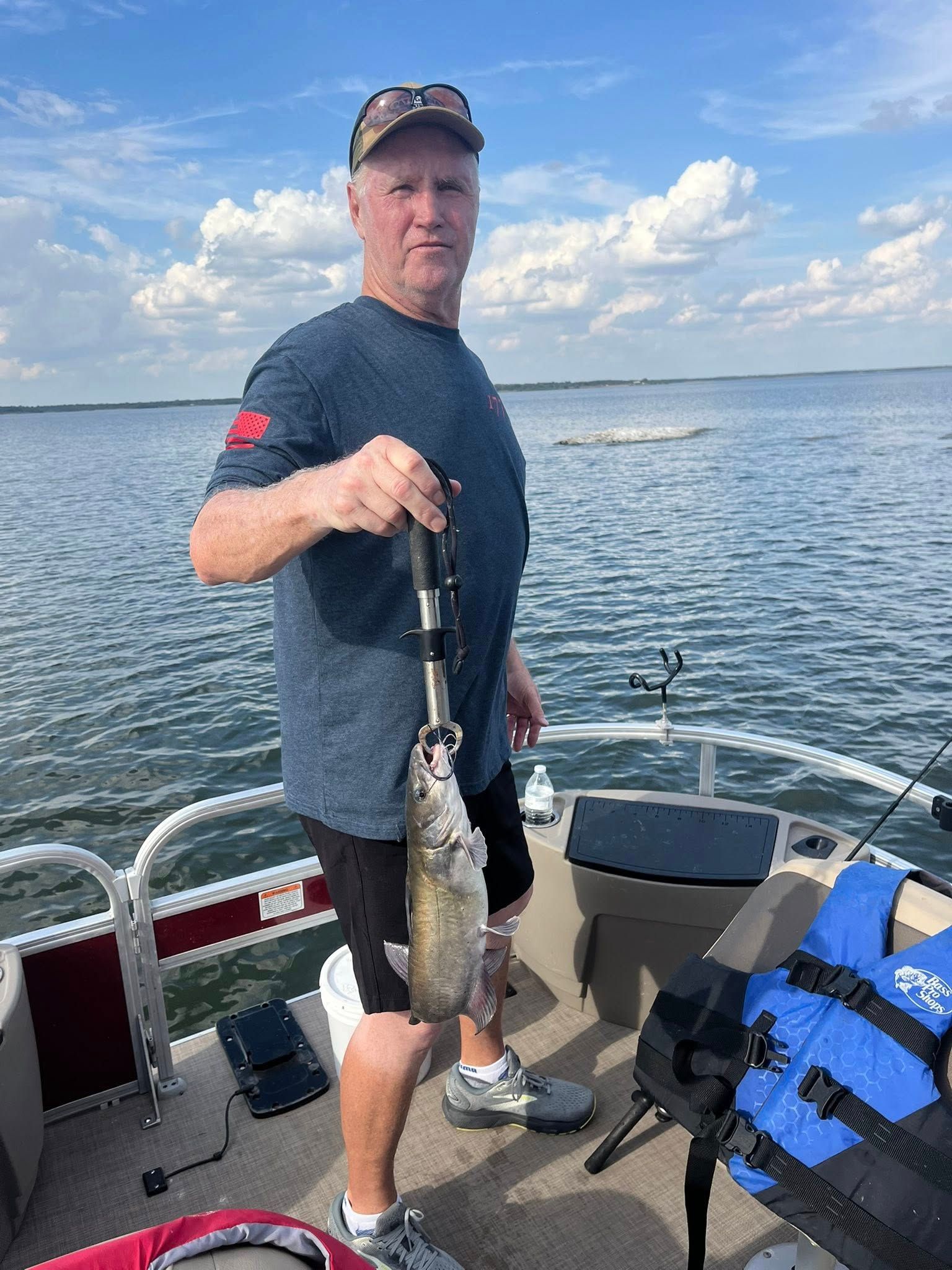 Man on a boat holds up a fish, lake in the background. Blue sky, wearing a cap, shirt, shorts.
