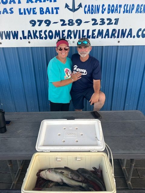 Couple poses with fish in a cooler at a marina, blue and white sign in background.
