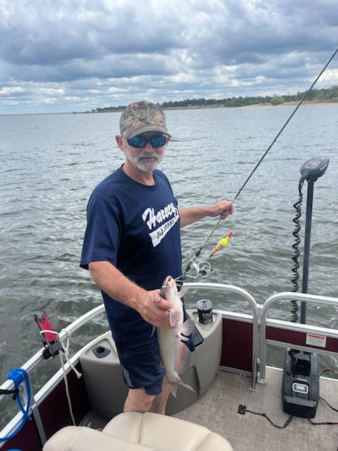 Man on a boat holds up a fish, fishing pole in hand, on a lake. Cloudy sky overhead.