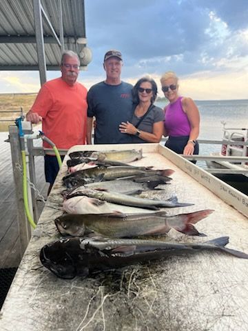 Four people stand behind a table with several catfish. The setting appears to be a dock near water.