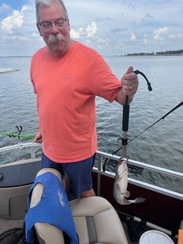 Man on boat, holding fish. Wearing orange shirt, blue shorts, water in background. Smiling.