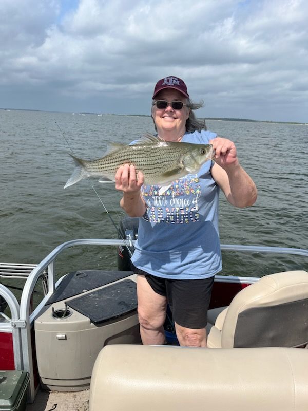 Woman on a boat holding a striped fish, smiling. Cloudy sky, water background.