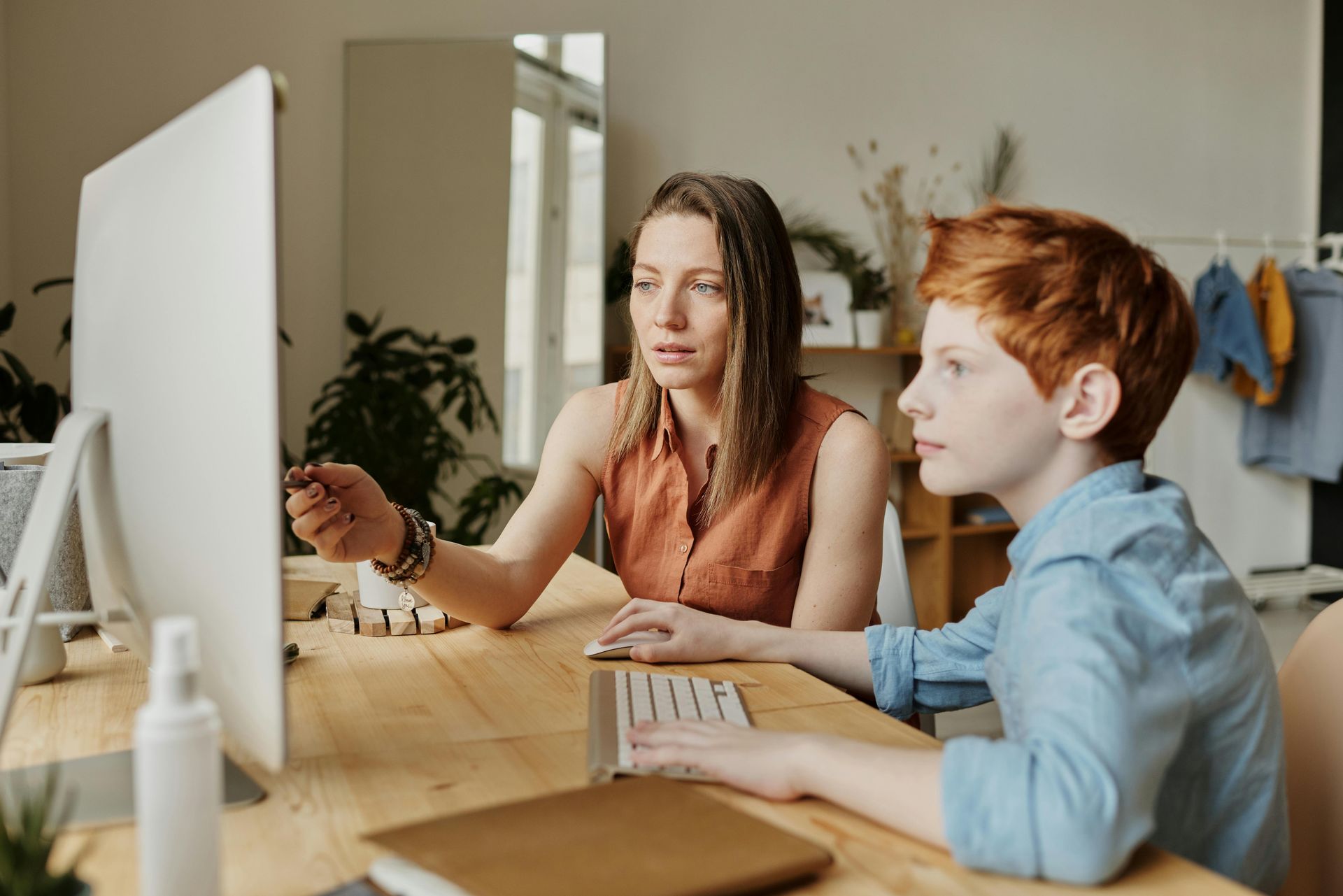 Woman points at computer screen, assisting a child with red hair. They sit at a wooden desk indoors.