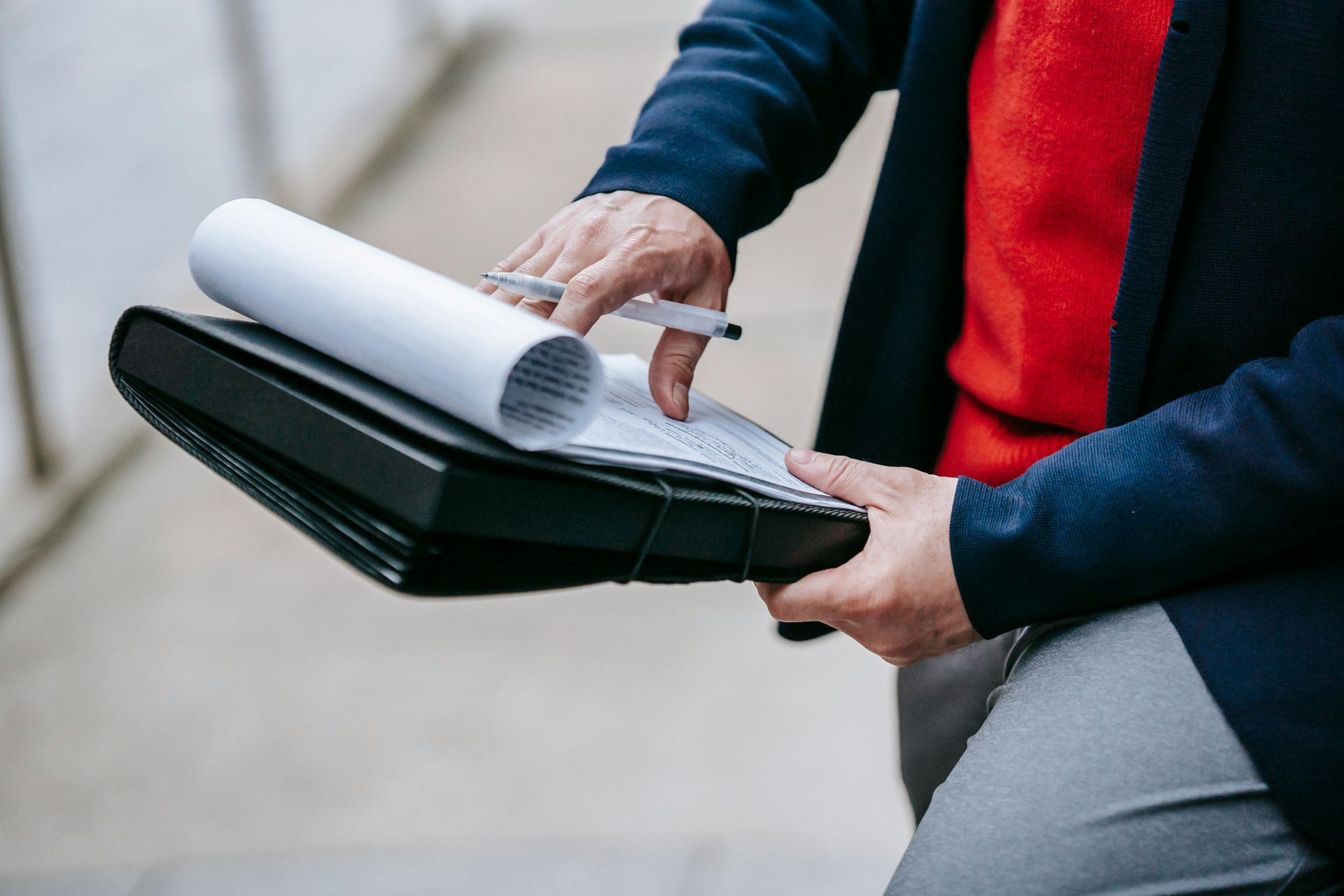 A person wearing a dark blue blazer and red shirt holds a black portfolio, pointing to a document with a silver pen.