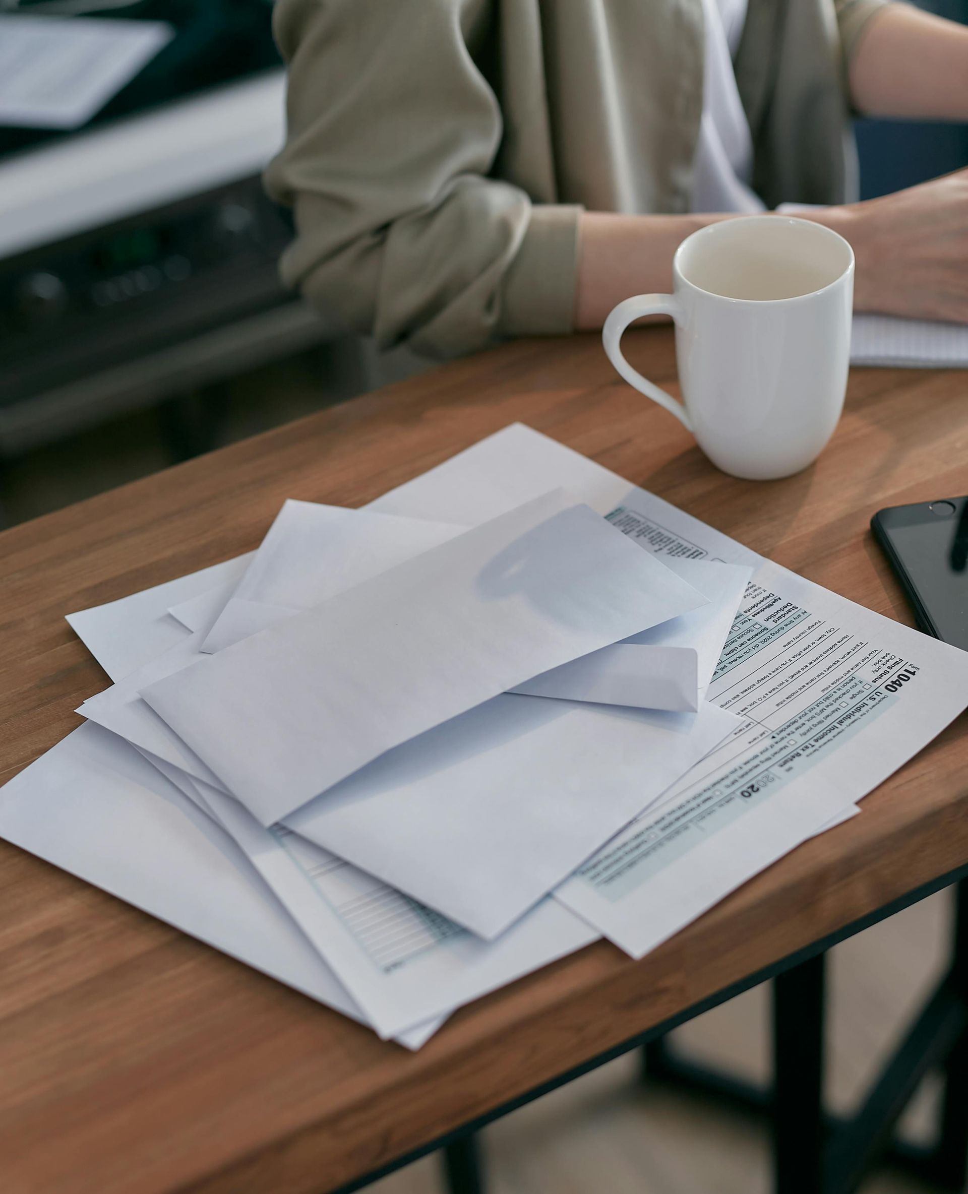 A person sits at a wooden desk with a white mug and a pile of documents and envelopes.