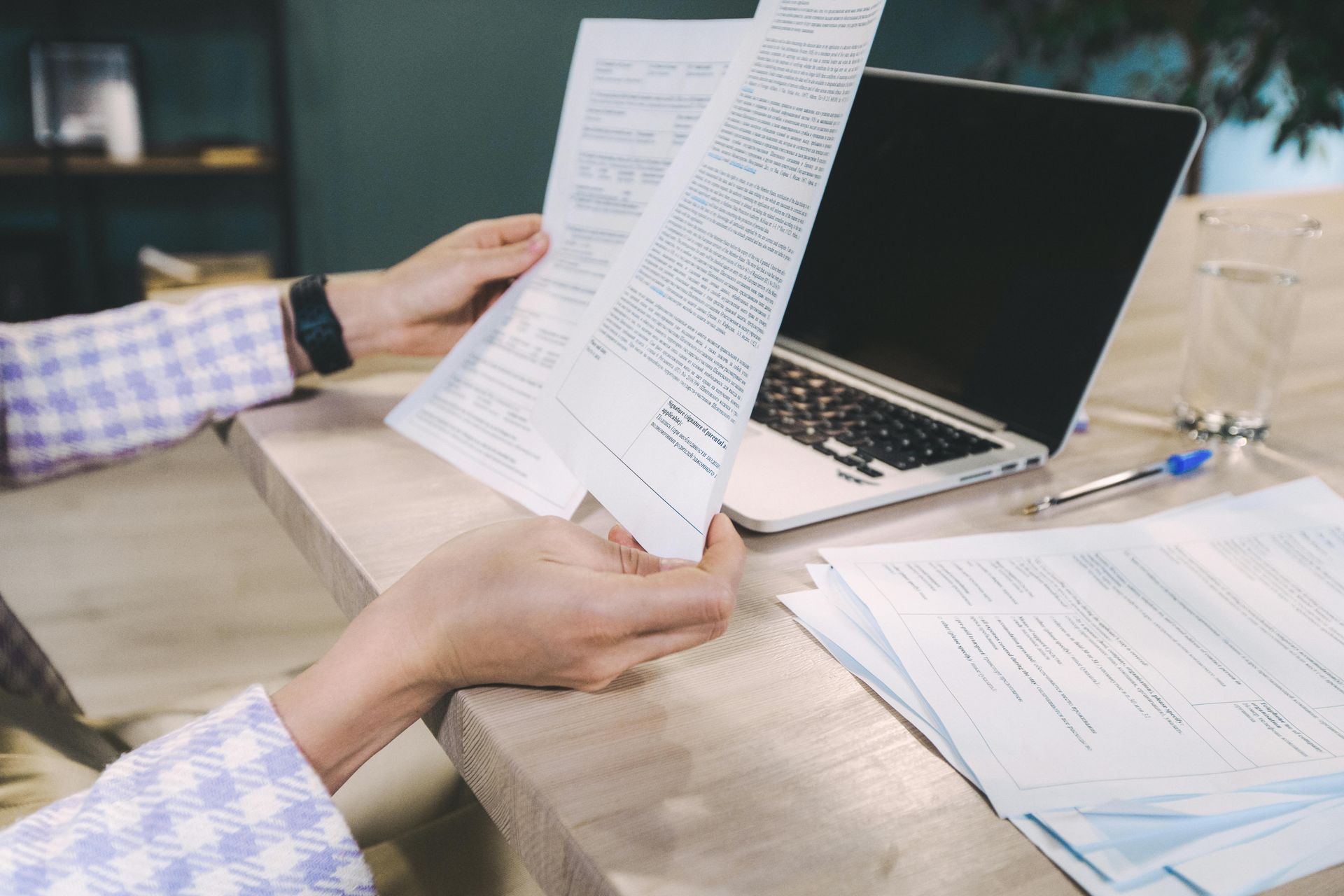 A person in a checkered shirt holds documents at a desk with a laptop, a glass of water, and a pen.