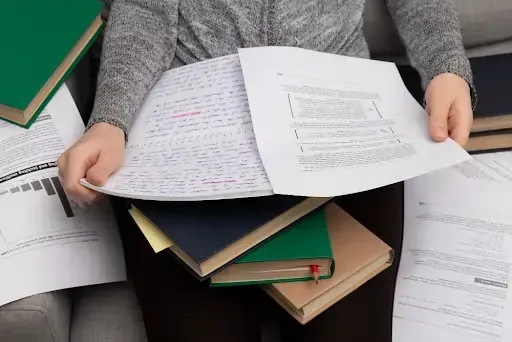 Person reviewing documents to shred in education, surrounded by books, handwritten notes, and printe
