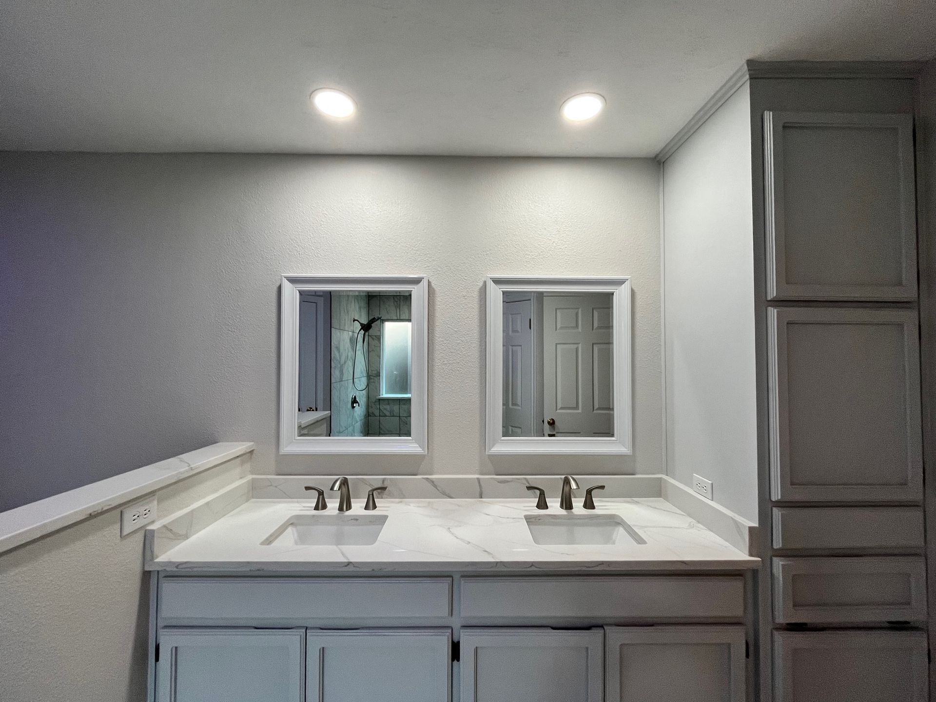 Bathroom with double sinks, white cabinets, mirrors, and a tall storage cabinet
