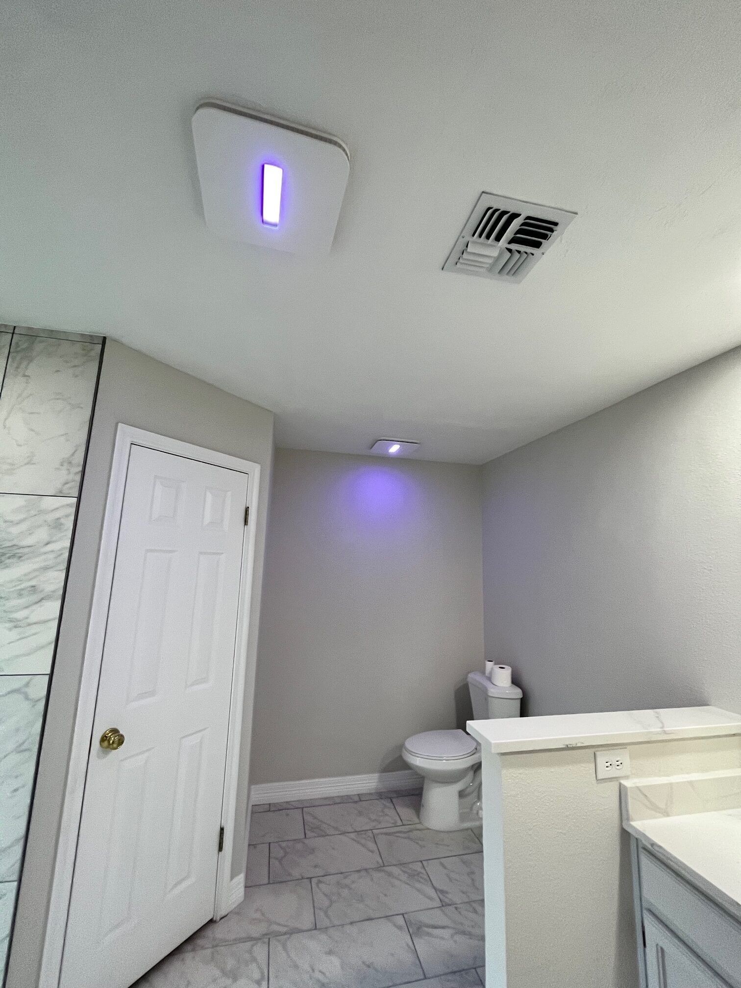 Bathroom with white door, toilet, gray walls, and a ceiling-mounted device emitting a purple light