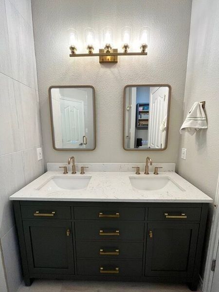 Bathroom with dark green vanity, white countertop, two mirrors, gold fixtures, and bright overhead lights