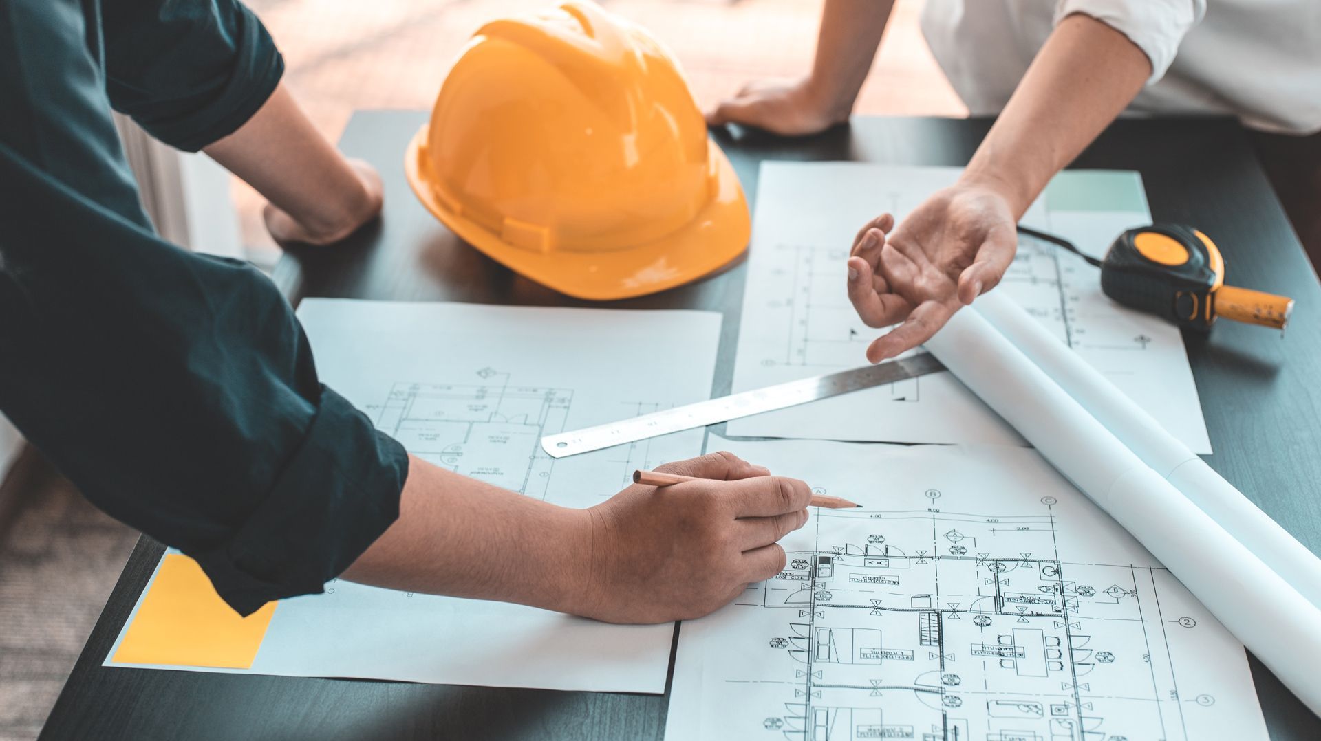 Two people reviewing blueprints, pointing; hard hat and measuring tape on the table