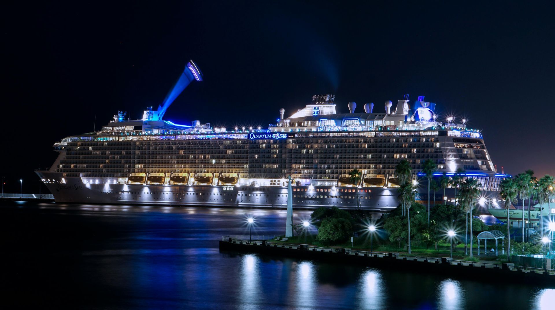 Large cruise ship illuminated at night docked in a harbor, blue and white lights reflecting on the water.