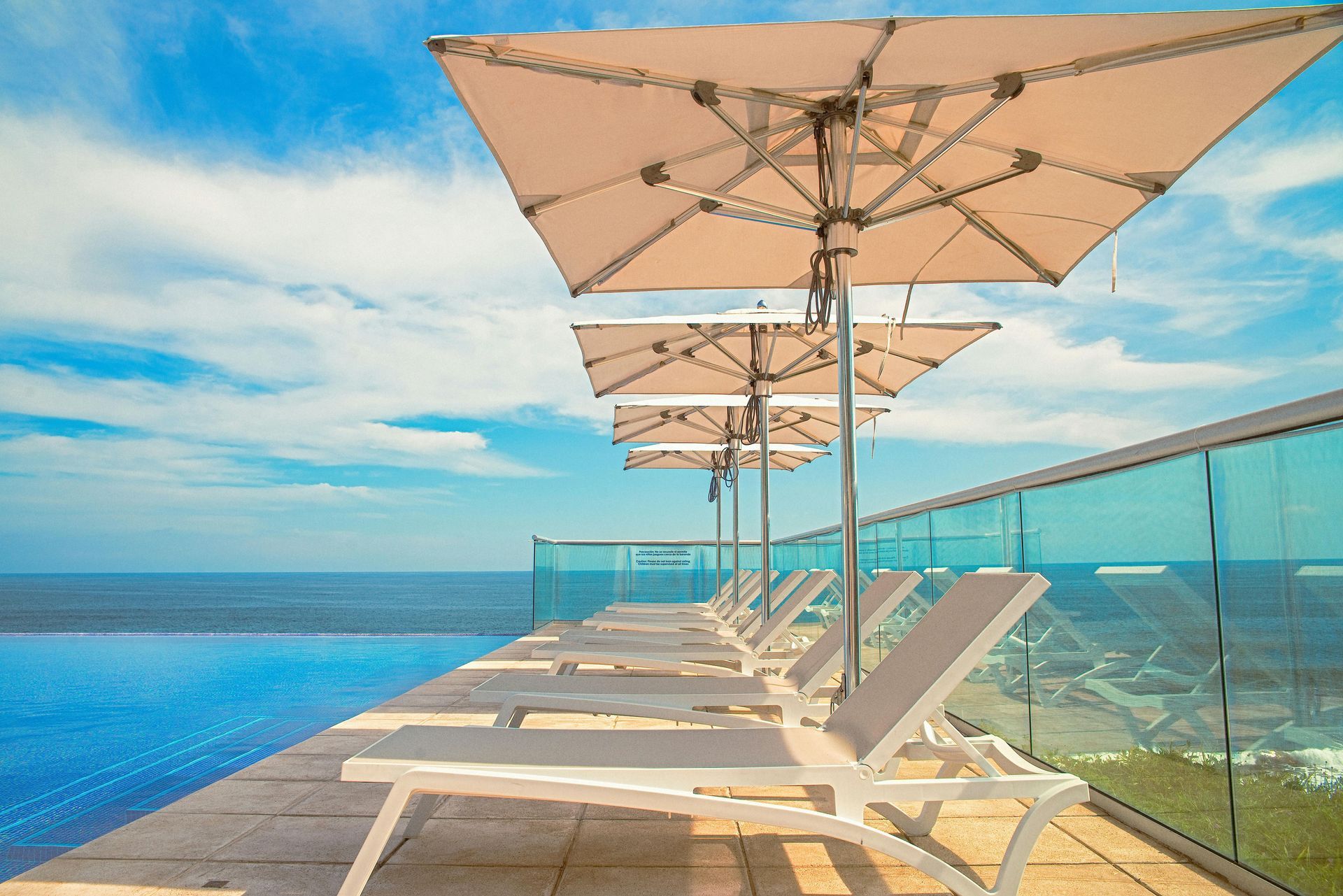 Lounge chairs under umbrellas beside a blue infinity pool, overlooking the ocean under a cloudy sky.