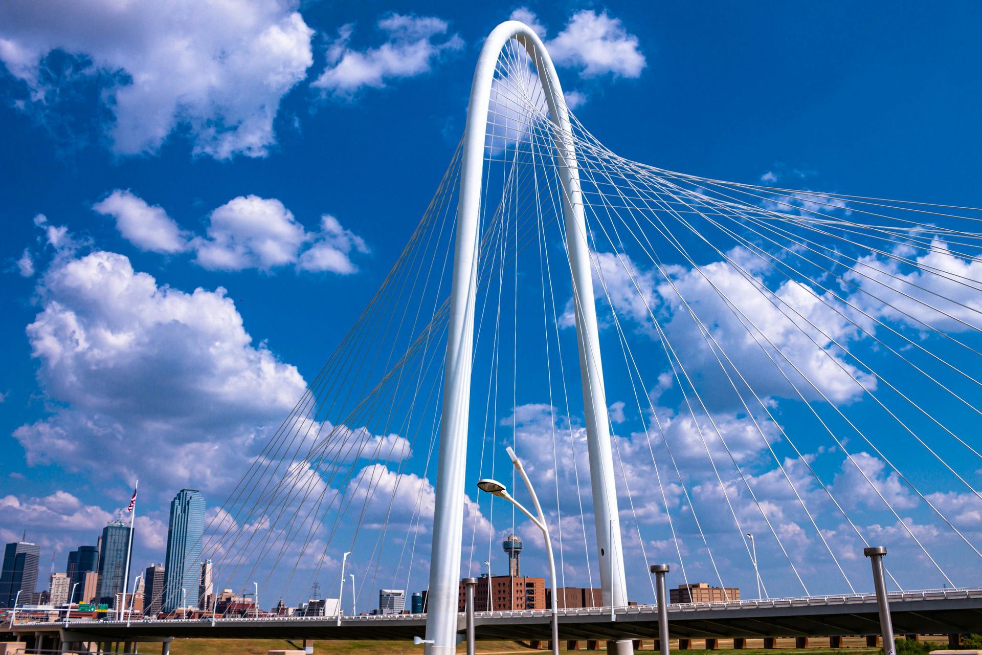 White arch suspension bridge over a road; Dallas skyline in background, blue sky with clouds.
