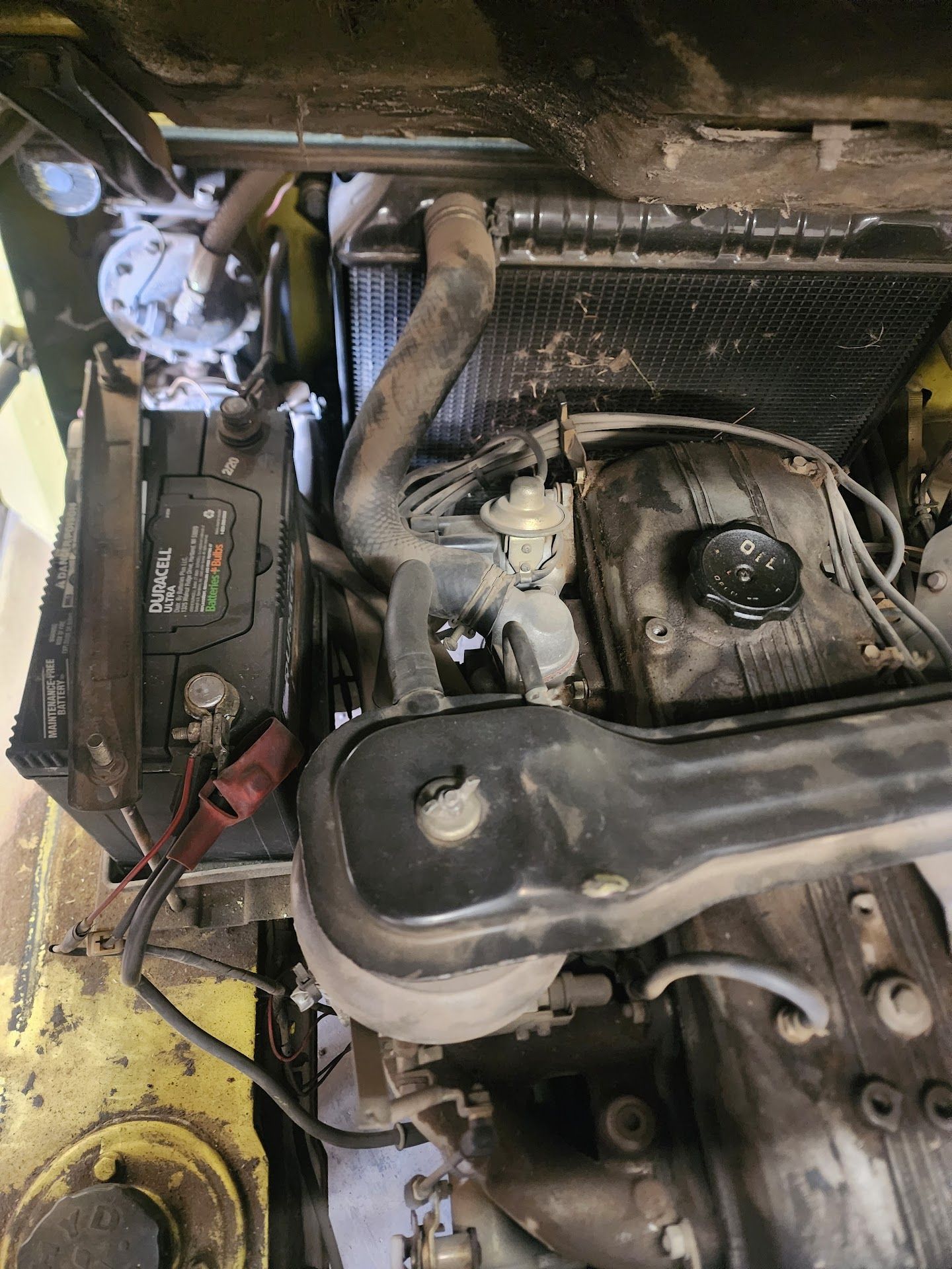 Battery and engine components of a yellow car, including radiator, air filter, and hoses. | TST Mechanic Services