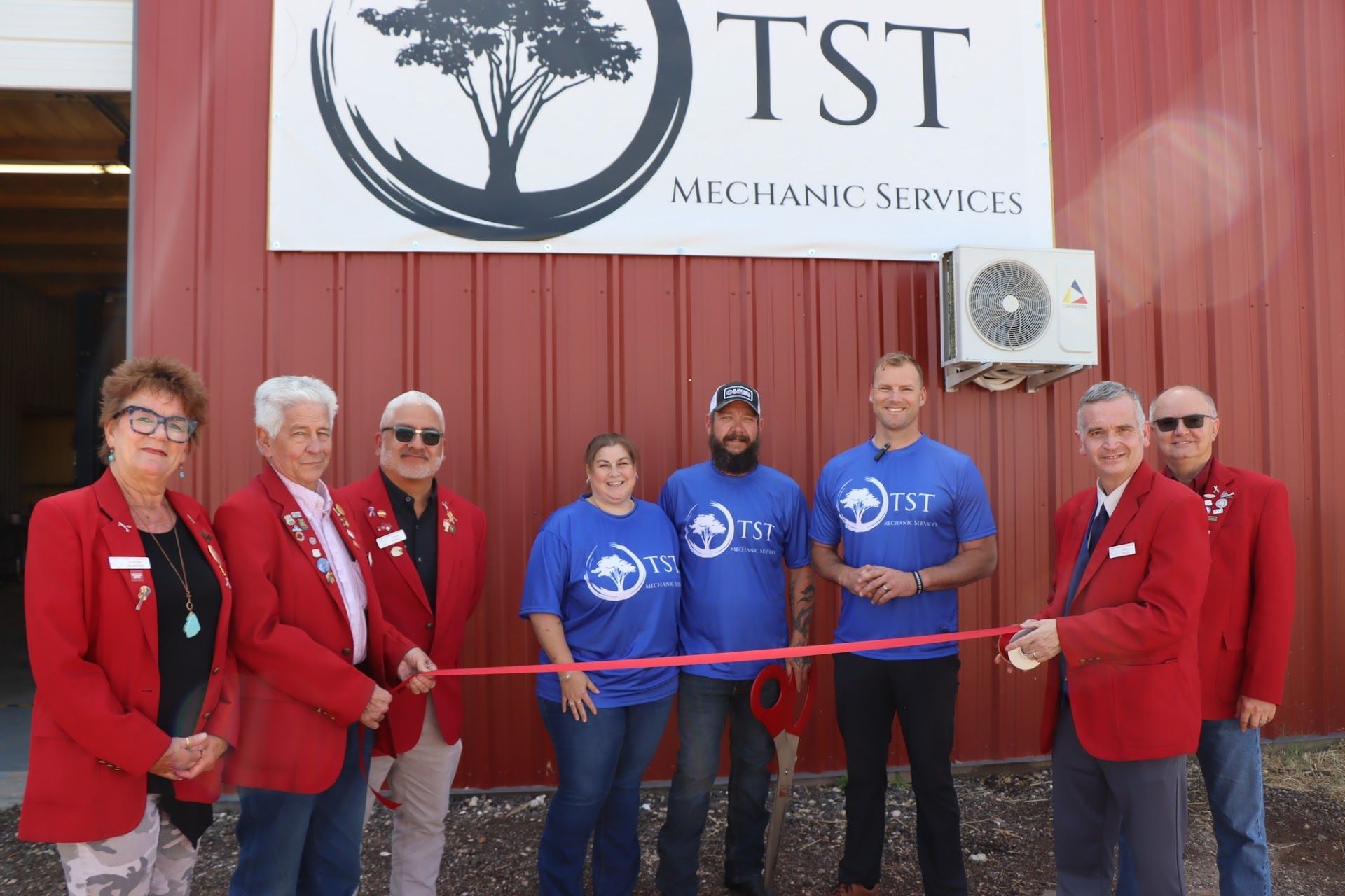 People in red jackets and blue shirts cut a ribbon in front of a business sign,